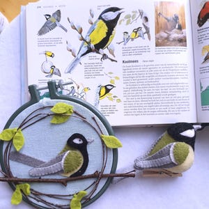 In this picture:  hand made wall decoration with a felt birds on linen in an embroidery hoop, the bird is a Great tit. The hoop is lying besides a book with a picture of the Great tit and a hand made felt Great tit in real life size on a white surface. This easy to make wall decoration is made with a PDF hand sewing pattern from WoolFeltWonders by Tintangel.