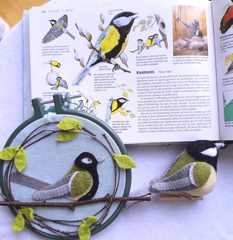 In this picture:  hand made wall decoration with a felt birds on linen in an embroidery hoop, the bird is a Great tit. The hoop is lying besides a book with a picture of the Great tit and a hand made felt Great tit in real life size on a white surface. This easy to make wall decoration is made with a PDF hand sewing pattern from WoolFeltWonders by Tintangel.