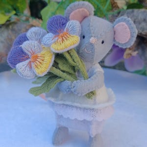 Op de afbeelding: Een handgemaakte vilten muisfiguur met een boeket geborduurde viooltjes. De muis is lichtgrijs met roze accenten op de oren en neus. De viooltjes zijn paars, geel en blauw met groene stelen. De muis draagt een witte en lavendelkleurige jurk.