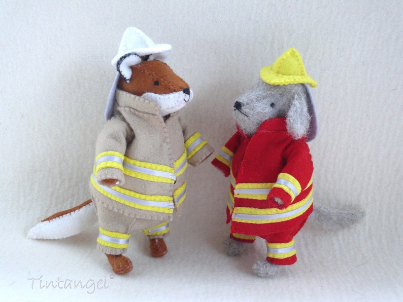 In this picture: two handmade felt toy animals, fox and dog, both dressed as a firefighter, one in a red costume, the other in a sand-colored costume. All clothing has refelcting stripes in yellow and silver. The dog with the red costume wears a yellow helmet, the other has a white helmet. All is made with a PDF handsewing pattern from WoolFeltWonders, part of a series with toy animal and clothing patterns to mix and match, to portray hobby or profession for play.