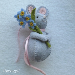 May include: A grey felt mouse holding a bouquet of blue forget-me-not flowers. The mouse has pink ears and a pink ribbon around its neck. The mouse is holding the flowers in its paws.