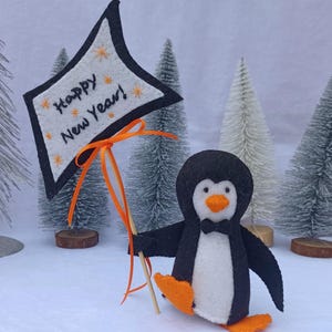 以下が含まれることがあります： 「Happy New Year！」と書かれた看板を持つ、手作りのフェルトペンギンの置物。 ペンギンは黒と白で、オレンジ色の足とくちばしがあり、蝶ネクタイをしています。 看板は白で、黒い縁取りとオレンジ色の文字が施されています。