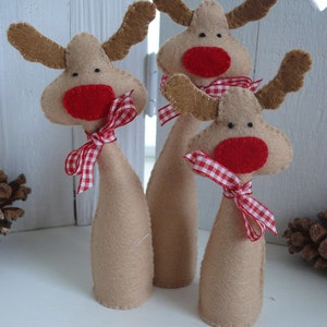 Peut inclure: Trois décorations de rennes en feutre marron avec des nez en feutre rouge et des nœuds en ruban vichy blanc et rouge. Les rennes sont debout sur une surface blanche.