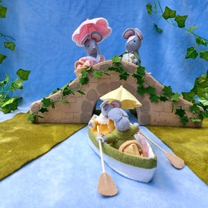 Può includere: Un bizzarro diorama in feltro con figure di topi. Due topi sono in piedi su un ponte beige adornato con viti verdi, mentre altri remano su una barca sotto un ombrello giallo. La scena è ambientata su uno sfondo blu con feltro verde e giallo che rappresenta l'acqua e la terra.
