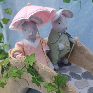 In this picture: a felt handmade  bridge, old style, with ivy vines along the upper side. The bridge is decorated with a stone pattern and is standing on two green waterfronts above blue water. This bridge is part of a Summer sewing project with elegant mice dressed in vintage clothing, Father, mother and two children in a boat.  In this picture the parents dressed in pink and green with an umbrella en walking stick.  All made of wool felt with a PDF hand sewing pattern from WoolFeltwonders.