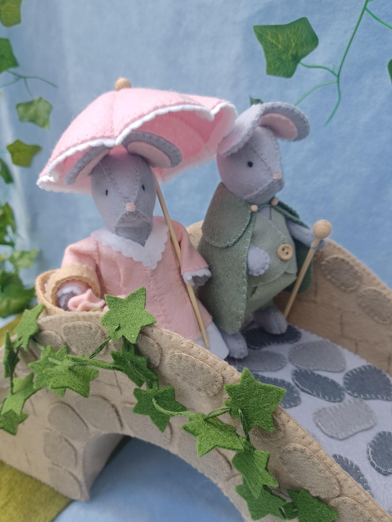 In this picture: a felt handmade  bridge, old style, with ivy vines along the upper side. The bridge is decorated with a stone pattern and is standing on two green waterfronts above blue water. This bridge is part of a Summer sewing project with elegant mice dressed in vintage clothing, Father, mother and two children in a boat.  In this picture the parents dressed in pink and green with an umbrella en walking stick.  All made of wool felt with a PDF hand sewing pattern from WoolFeltwonders.