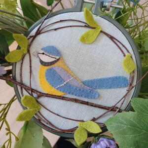 In this picture:  hand made wall decoration with a felt birds on linen in an embroidery hoop, the bird is a blue tit. The hoop is lying between flowers and leaves. This easy to make wall decoration is made with a PDF hand sewing pattern from WoolFeltWonders by Tintangel.