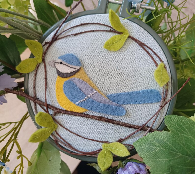 In this picture:  hand made wall decoration with a felt birds on linen in an embroidery hoop, the bird is a blue tit. The hoop is lying between flowers and leaves. This easy to make wall decoration is made with a PDF hand sewing pattern from WoolFeltWonders by Tintangel.