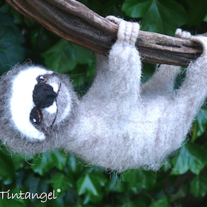 May include: A gray and white sloth with black eyes hanging from a branch. The sloth is made of felt and has a whimsical expression.