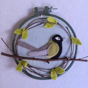 In this picture:  hand made wall decoration with a felt birds on linen in an embroidery hoop, the bird is a Great tit. The hoop is lying on a white surface. These easy to make wall decoration is made with a PDF hand sewing pattern from WoolFeltWonders by Tintangel.