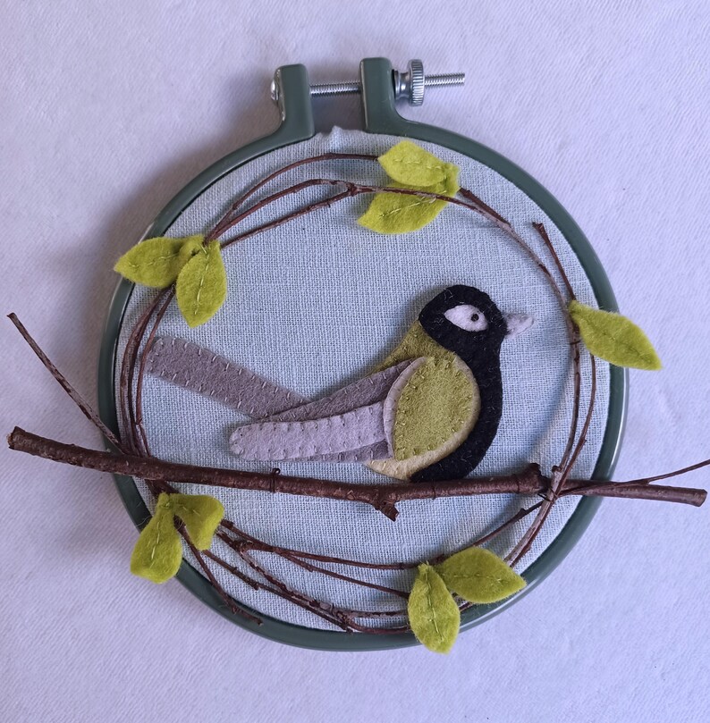 In this picture:  hand made wall decoration with a felt birds on linen in an embroidery hoop, the bird is a Great tit. The hoop is lying on a white surface. These easy to make wall decoration is made with a PDF hand sewing pattern from WoolFeltWonders by Tintangel.