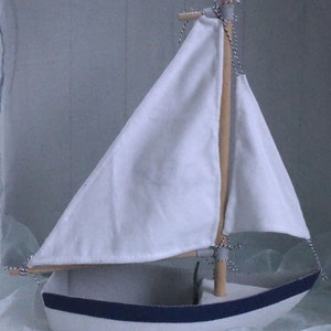 In this picture: a handmade sailing boat, made from felt in white with soft grey and dark blue, with a dark blue steering wheel. The sail is white with bleu lettern WFW.The boat is standing on en soft bleu surface. This boat is made with a PDF handsewing pattern from WoolFeltWonders and is part of a series with felt toy animals ad costumes to portray a profession or hobby. This boat is lovely in combination with some toy animals and the sailor clothing, also a PDF pattern in this series.