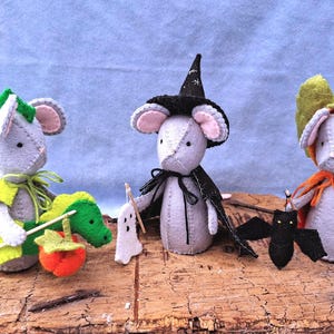 Puede incluir: Tres figuras de ratones de fieltro hechas a mano disfrazadas para Halloween. El ratón de la izquierda lleva un disfraz de dragón verde con una calabaza. El central lleva un sombrero de bruja negro y capa con un fantasma. El de la derecha lleva una capa naranja con un murciélago.