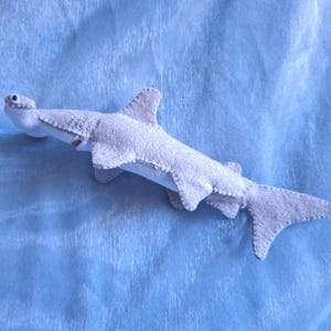 May include: A light gray felt hammerhead shark plush toy featuring visible stitched seams along the edges, a white underbelly, and small black bead eyes. The shark is oriented horizontally with its wide head, dorsal fin, and tail clearly visible.