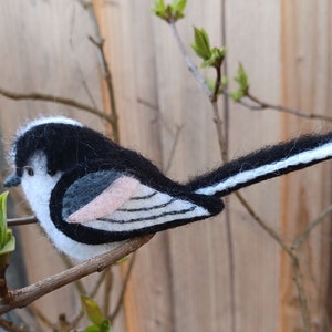 Op de afbeelding: Een handgevoelde langstaartmees van zwart, wit en roze vilt, die op een tak zit.