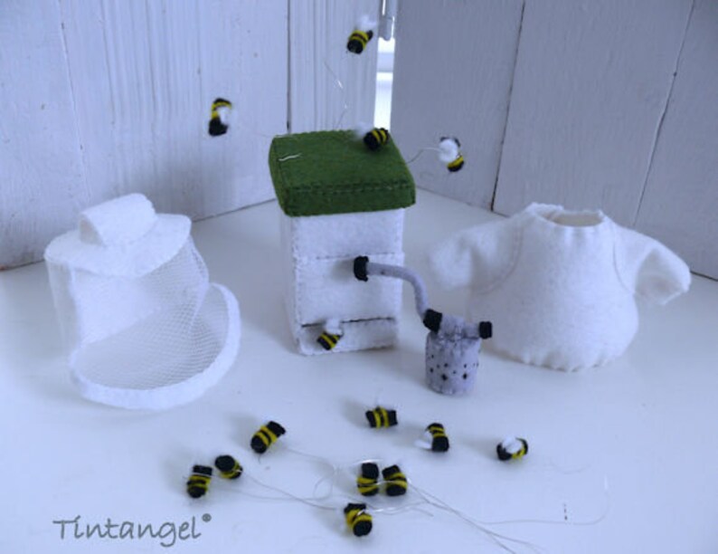 In this picture: a handmade set with clothing and accessoiries to dress a felt toy animal as a Beekeeper, with white jacket, bee hood, beehive, smoke pipe and many bees. All this is is made with PDF hand sewing pattern from WoolFeltWonders by Tintangel and is part of a series with felt toy animals and all kinds of clothing sets to mix and match to portray hobby or profession. Easy to make animals and clothing sets for decoration and play.