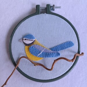 In this picture:  hand made wall decoration with a felt birds on linen in an embroidery hoop, the bird is a blue tit. The hoop is lying on a white surface. These easy to make wall decoration is made with a PDF hand sewing pattern from WoolFeltWonders by Tintangel.