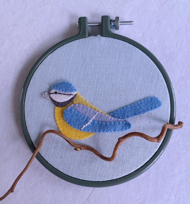 In this picture:  hand made wall decoration with a felt birds on linen in an embroidery hoop, the bird is a blue tit. The hoop is lying on a white surface. These easy to make wall decoration is made with a PDF hand sewing pattern from WoolFeltWonders by Tintangel.