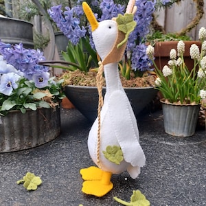 May include: A white felt goose with yellow feet and a brown felt hat with a green felt shamrock. The goose is standing on a gray surface with green felt shamrocks scattered around it.