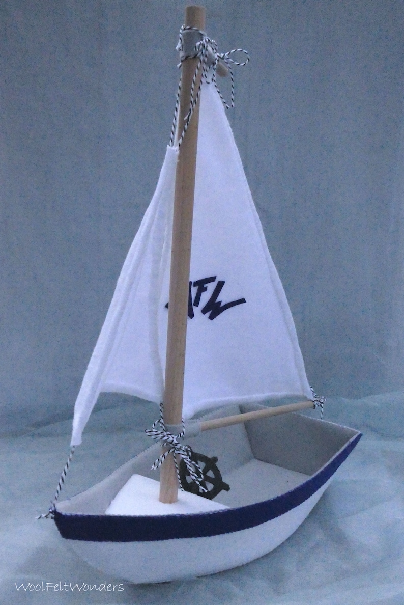 In this picture: a handmade sailing boat, made from felt in white with soft grey and dark blue, with a dark blue steering wheel. The sail is white with bleu lettern WFW.The boat is standing on en soft bleu surface. This boat is made with a PDF handsewing pattern from WoolFeltWonders and is part of a series with felt toy animals ad costumes to portray a profession or hobby. This boat is lovely in combination with some toy animals and the sailor clothing, also a PDF pattern in this series.