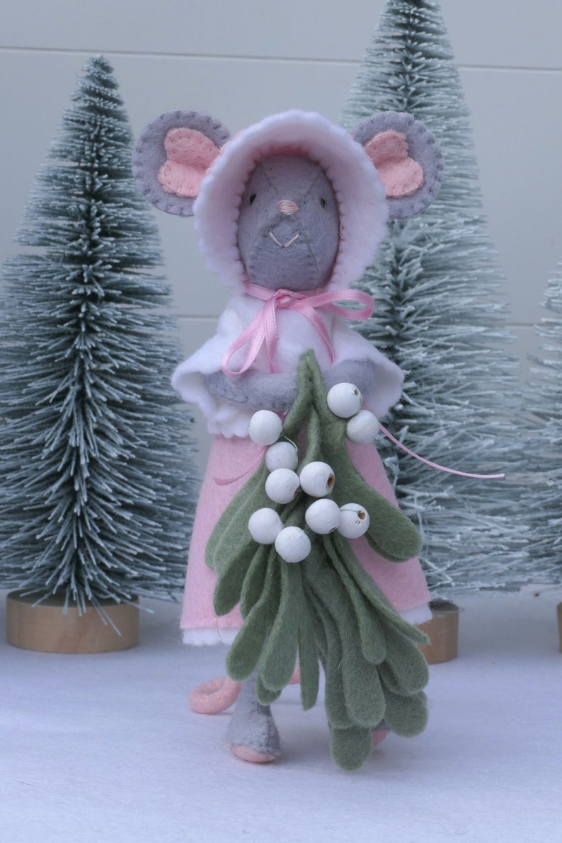 In this picture: a sweet handmade felt mouse with pink dress, white cape and pink and white bonnet, holding a bunch of felt mistletoe with white berries between her hands. She is standing on front of two fake trees on a white surface. This sweet grey mouse is made with a PDF hand sewing pattern from WoolFeltWonders by Tintangel.