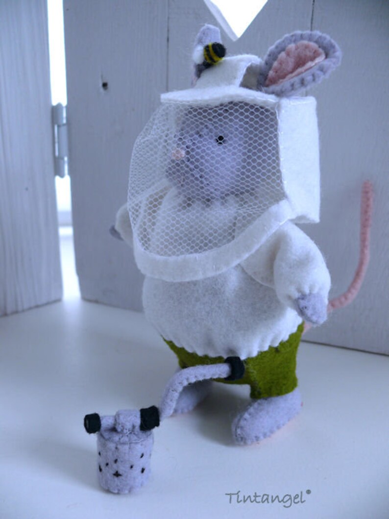 In this picture: a handmade felt mouse dressed as a beekeeper, with white jacket, bee hood, and smoke pipe. All this is is made with PDF hand sewing pattern from WoolFeltWonders by Tintangel and is part of a series with felt toy animals and all kinds of clothing sets to mix and match to portray hobby or profession. Easy to make animals and clothing sets for decoration and play.