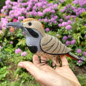 May include: A handmade felt bird with a tan body, gray wings, and black and gray accents. The bird has a gray beak and is perched on a hand. The background features pink flowers and green foliage.