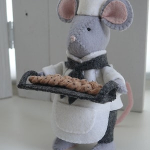 Könnte beinhalten: Eine graue Filzmaus mit einer weißen Kochmütze und einer Schürze hält ein Backblech mit braunen Brötchen.