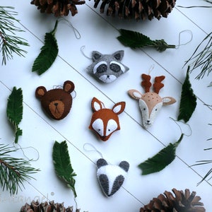 Könnte beinhalten: Fünf Filz-Tier-Anhänger, darunter ein Waschbär, ein Bär, ein Fuchs, ein Reh und ein Panda, die an einer weißen Schnur an einem weißen Holzgrund hängen. Die Anhänger sind von Kiefernästen und Tannenzapfen umgeben.