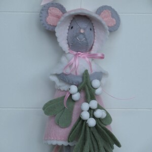 In this picture: a sweet handmade felt mouse with pink dress, white cape and pink and white bonnet, holding a bunch of felt mistletoe with white berries between her hands. She is hanging in a wreath with pink bow on a white surface. This sweet grey mouse is made with a PDF hand sewing pattern from WoolFeltWonders by Tintangel.