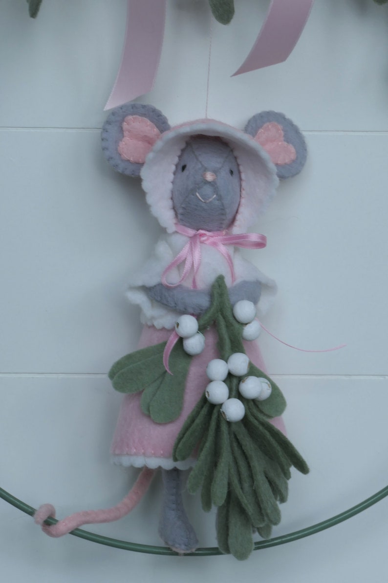 In this picture: a sweet handmade felt mouse with pink dress, white cape and pink and white bonnet, holding a bunch of felt mistletoe with white berries between her hands. She is hanging in a wreath with pink bow on a white surface. This sweet grey mouse is made with a PDF hand sewing pattern from WoolFeltWonders by Tintangel.