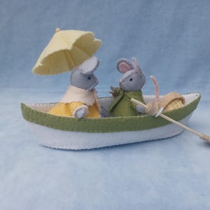 Peut inclure: Un bateau en feutre fait à la main avec deux souris grises vêtues de tenues jaunes et vertes. Une souris tient un parasol jaune. Le bateau a une coque blanche avec une bande verte et une rame en bois. Le décor est sur fond bleu clair.