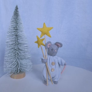 May include: A gray felt mouse wearing a light blue onesie holds three yellow felt stars on wooden sticks. The mouse is standing on a white surface in front of a white flocked Christmas tree.