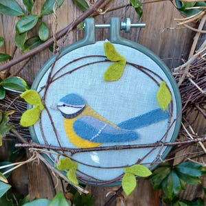 In this picture:  hand made wall decoration with a felt birds on linen in an embroidery hoop, the bird is a blue tit. The hoop is lying between  leaves in front of a wooden wall. This easy to make wall decoration is made with a PDF hand sewing pattern from WoolFeltWonders by Tintangel.
