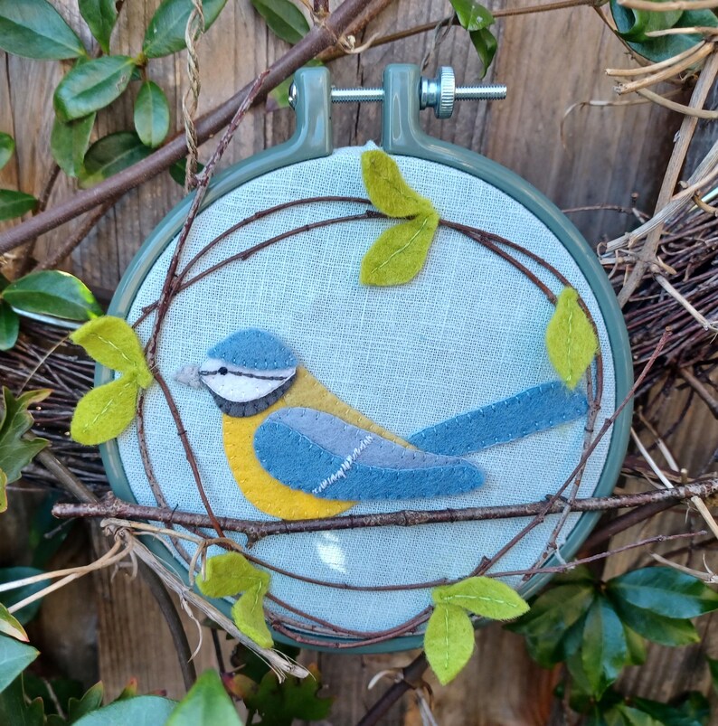 In this picture:  hand made wall decoration with a felt birds on linen in an embroidery hoop, the bird is a blue tit. The hoop is lying between  leaves in front of a wooden wall. This easy to make wall decoration is made with a PDF hand sewing pattern from WoolFeltWonders by Tintangel.
