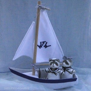 In this picture: a handmade sailing boat, made from felt in white with soft grey and dark blue, with a dark blue steering wheel. The sail is white with bleu lettern WFW. In  the boat two felt toy racoons in sailor costumes. This boat, the racoons and their clothing is made with a PDF handsewing pattern from WoolFeltWonders and is part of a series with felt toy animals ad costumes to portray a profession or hobby.