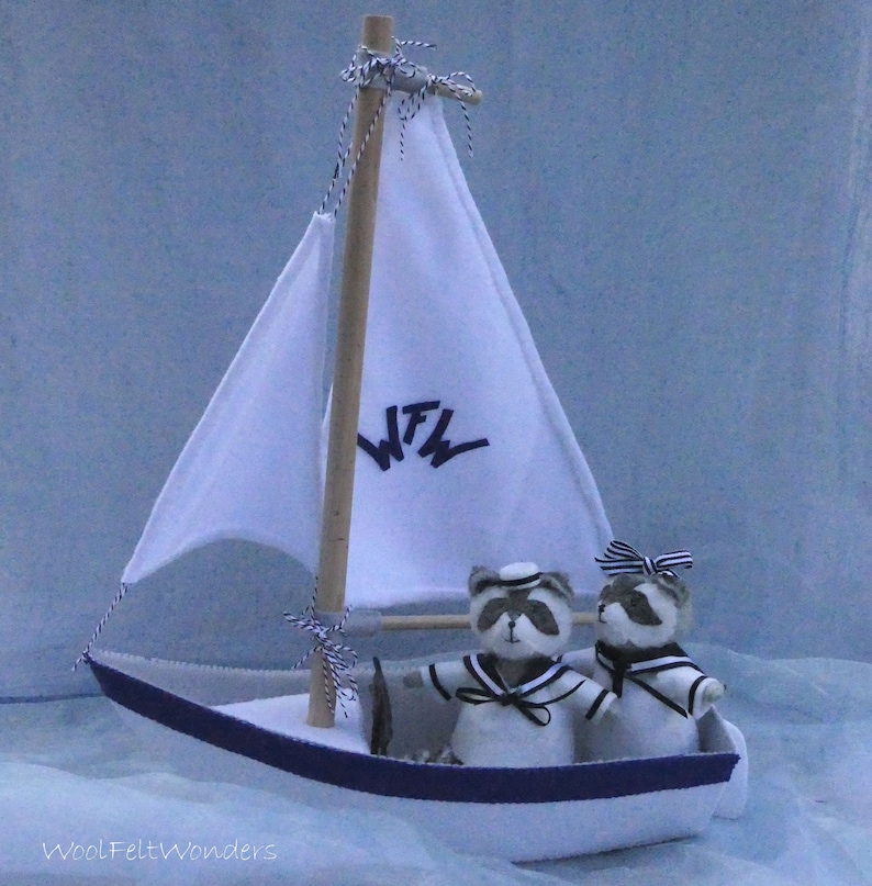 In this picture: a handmade sailing boat, made from felt in white with soft grey and dark blue, with a dark blue steering wheel. The sail is white with bleu lettern WFW. In  the boat two felt toy racoons in sailor costumes. This boat, the racoons and their clothing is made with a PDF handsewing pattern from WoolFeltWonders and is part of a series with felt toy animals ad costumes to portray a profession or hobby.