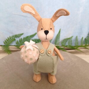May include: A felt bunny toy in tan and light green overalls, holding a pink bag with white spots and a white bow. The bunny has long ears and a black nose. The background is light blue with green fern leaves. A charming Easter decoration.