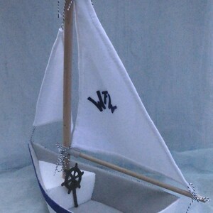 In this picture: a handmade sailing boat, made from felt in white with soft grey and dark blue, with a dark blue steering wheel. The sail is white with bleu lettern WFW.The boat is standing on en soft bleu surface. This boat is made with a PDF handsewing pattern from WoolFeltWonders and is part of a series with felt toy animals ad costumes to portray a profession or hobby. This boat is lovely in combination with some toy animals and the sailor clothing, also a PDF pattern in this series.