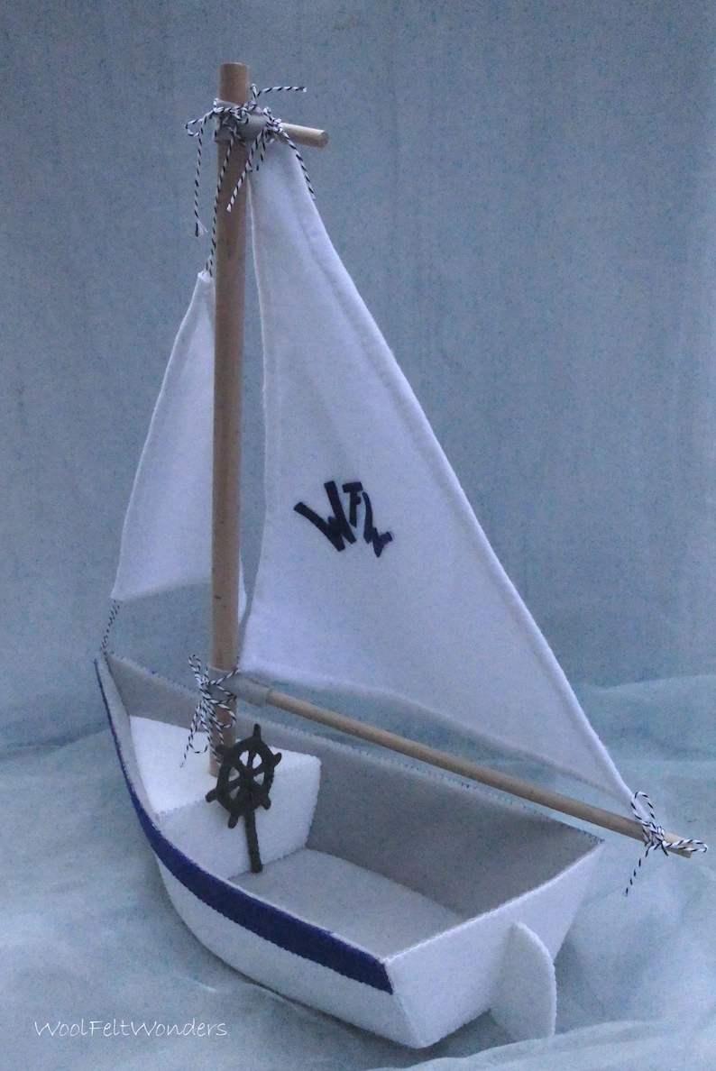 In this picture: a handmade sailing boat, made from felt in white with soft grey and dark blue, with a dark blue steering wheel. The sail is white with bleu lettern WFW.The boat is standing on en soft bleu surface. This boat is made with a PDF handsewing pattern from WoolFeltWonders and is part of a series with felt toy animals ad costumes to portray a profession or hobby. This boat is lovely in combination with some toy animals and the sailor clothing, also a PDF pattern in this series.