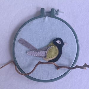 In this picture:  hand made wall decoration with a felt birds on linen in an embroidery hoop, the bird is a Great tit. The hoop is lying on a white surface. These easy to make wall decoration is made with a PDF hand sewing pattern from WoolFeltWonders by Tintangel.
