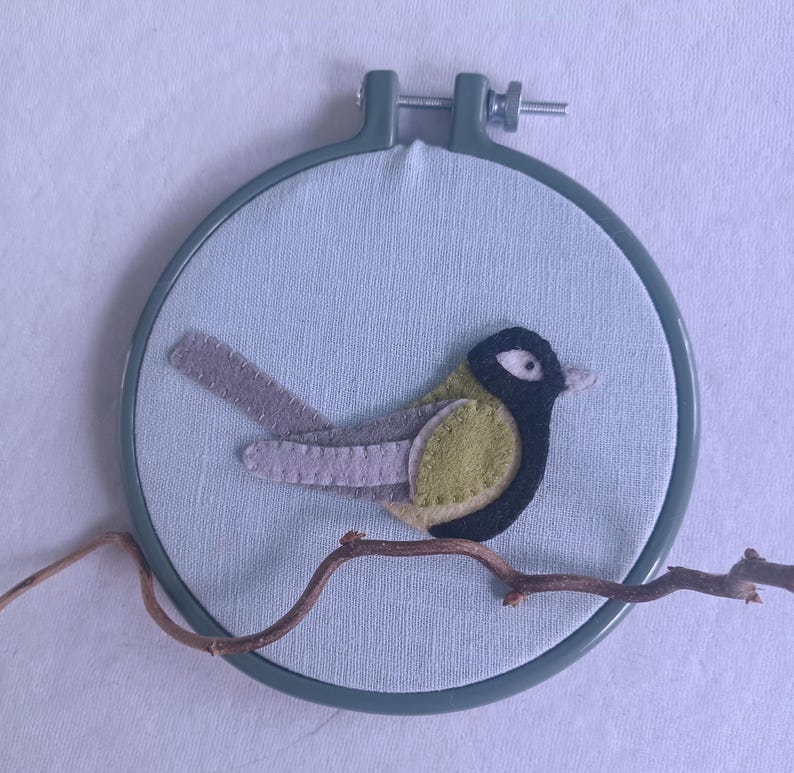 In this picture:  hand made wall decoration with a felt birds on linen in an embroidery hoop, the bird is a Great tit. The hoop is lying on a white surface. These easy to make wall decoration is made with a PDF hand sewing pattern from WoolFeltWonders by Tintangel.