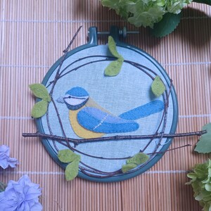 In this picture:  hand made wall decoration with a felt birds on linen in an embroidery hoop, the bird is a blue tit. The hoop is lying on a reed mat and some leaves above and besides it. These easy to make wall decoration is made with a PDF hand sewing pattern from WoolFeltWonders by Tintangel.