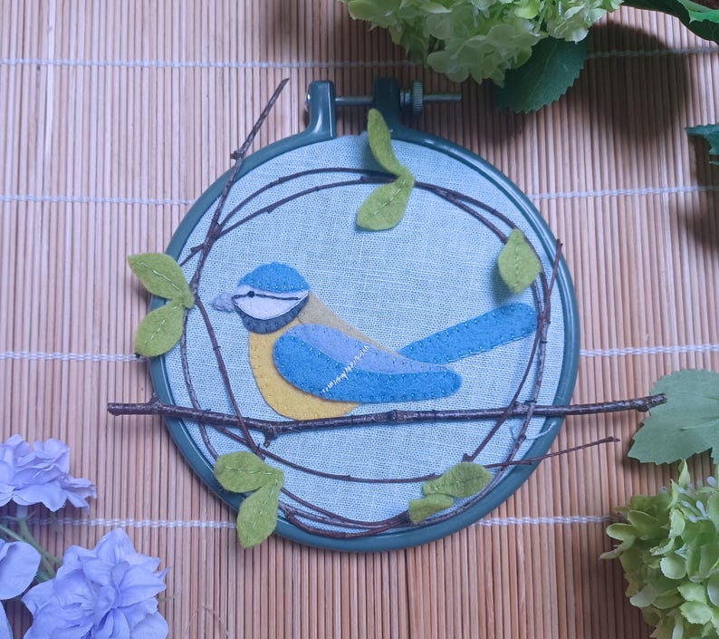 In this picture:  hand made wall decoration with a felt birds on linen in an embroidery hoop, the bird is a blue tit. The hoop is lying on a reed mat and some leaves above and besides it. These easy to make wall decoration is made with a PDF hand sewing pattern from WoolFeltWonders by Tintangel.