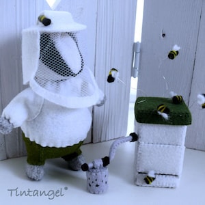 In this picture: a handmade badger dressed as a beekeeper, with white jacket, bee hood, beehive, smoke pipe and many bees. All this is is made with PDF hand sewing pattern from WoolFeltWonders by Tintangel and is part of a series with felt toy animals and all kinds of clothing sets to mix and match to portray hobby or profession. Easy to make animals and clothing sets for decoration and play.