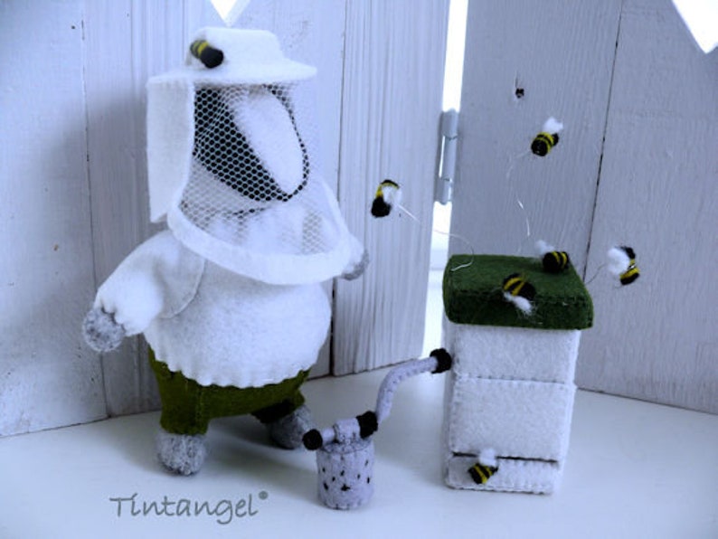 In this picture: a handmade badger dressed as a beekeeper, with white jacket, bee hood, beehive, smoke pipe and many bees. All this is is made with PDF hand sewing pattern from WoolFeltWonders by Tintangel and is part of a series with felt toy animals and all kinds of clothing sets to mix and match to portray hobby or profession. Easy to make animals and clothing sets for decoration and play.