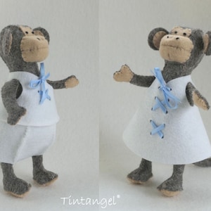 May include: A gray felt monkey wearing a white felt dress with blue ribbon accents. The monkey has a long tail and is standing on a white background.