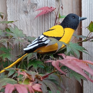May include: A felt bird with a black head, yellow body, and white and black striped wings perches on a leafy branch. The bird's tail is yellow, and it has a gray beak. The background includes a wooden fence and red and green leaves.