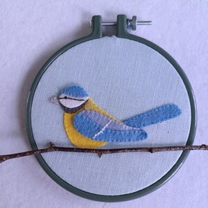 In this picture:  hand made wall decoration with a felt birds on linen in an embroidery hoop, the bird is a blue tit. The hoop is lying on a white surface. These easy to make wall decoration is made with a PDF hand sewing pattern from WoolFeltWonders by Tintangel.
