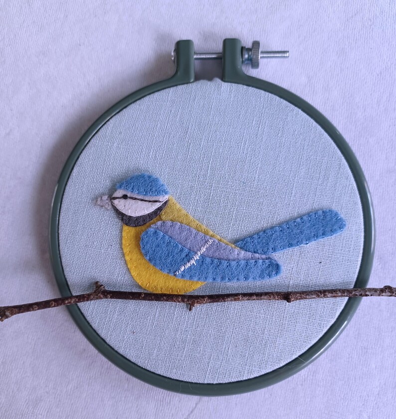 In this picture:  hand made wall decoration with a felt birds on linen in an embroidery hoop, the bird is a blue tit. The hoop is lying on a white surface. These easy to make wall decoration is made with a PDF hand sewing pattern from WoolFeltWonders by Tintangel.